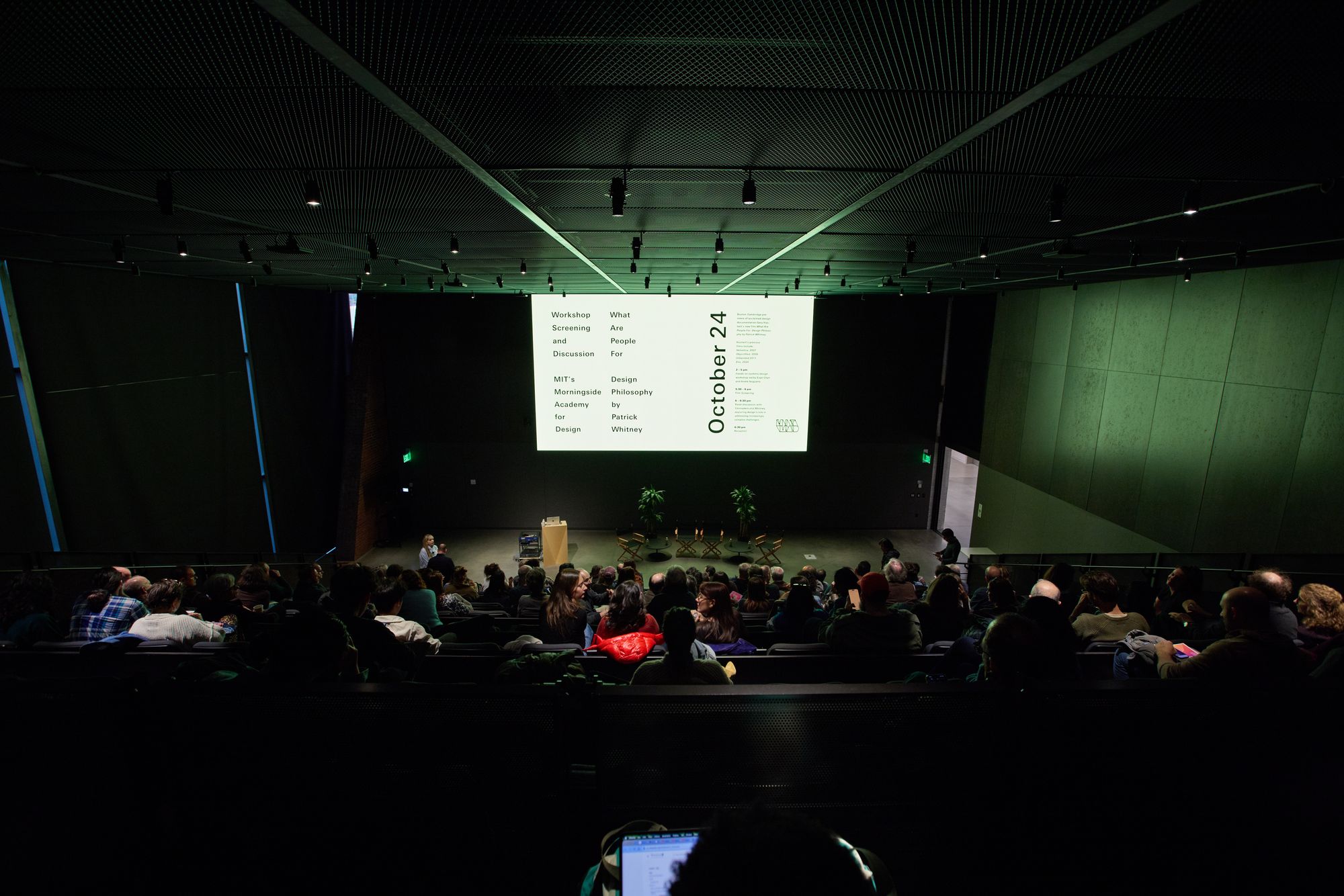 The view from above: A full auditorium, director's chairs set up for the panel, and the holding slide sharing the event name and date.